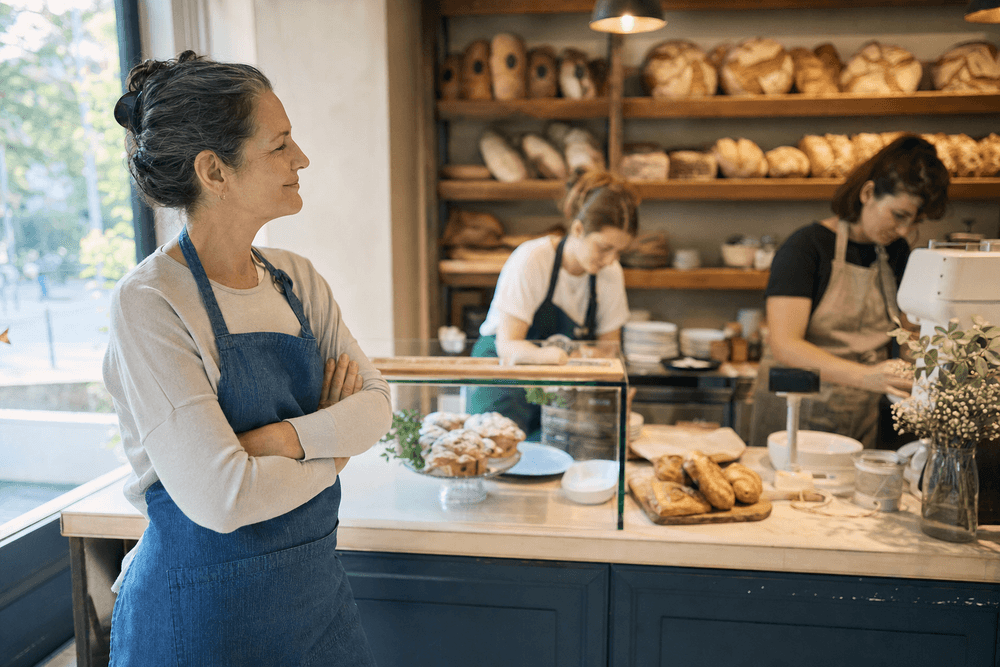 A calm bakery owner stepping back to watch two staff run the morning rush behind the counter.