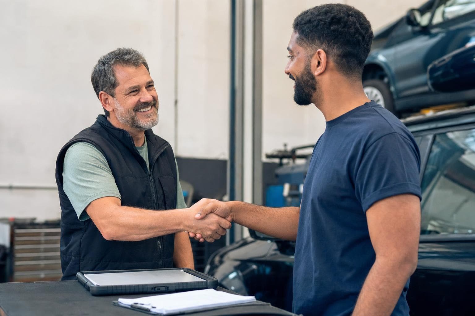 An auto-shop owner shaking a customer's hand in the bay while a tablet handles the paperwork.