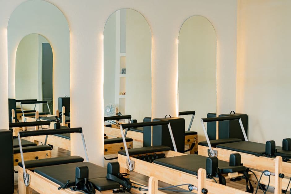 A bright, empty pilates studio with rows of unused reformer machines lit by warm light through arched mirrors, suggesting a quiet, emptying class.