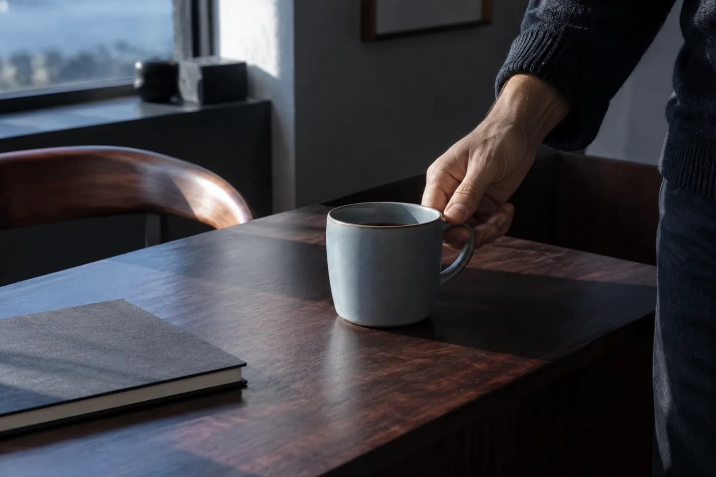 A hand setting a ceramic cup beside a closed notebook in morning light
