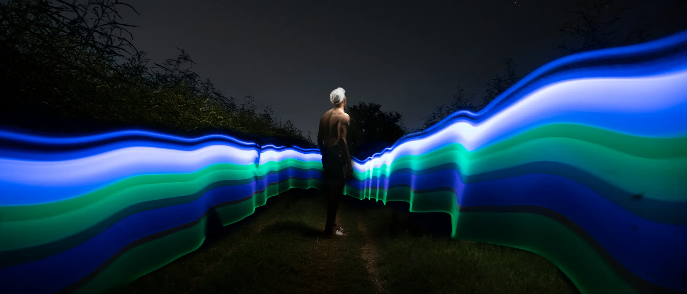 A lone figure at night between two long-exposure ribbons of blue and green light