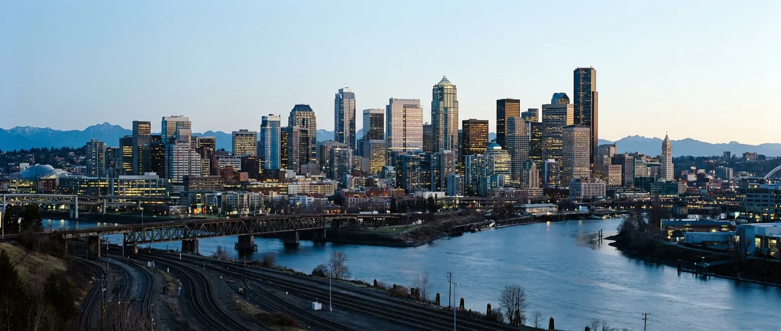 The downtown skyline at dusk seen across the river confluence and the rail yards.