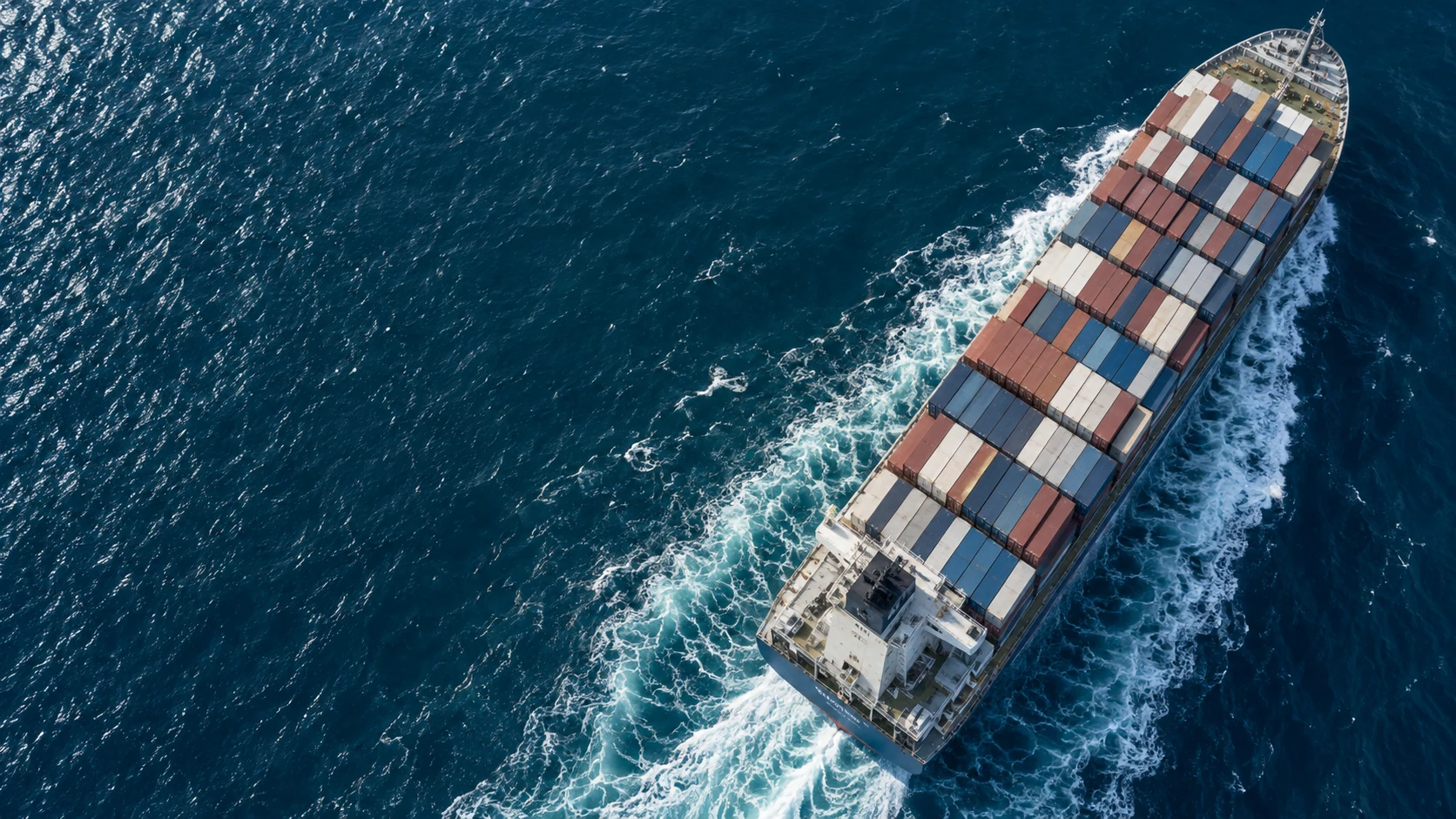 A container vessel carrying blue, ivory and burnt-red containers through open ocean water.