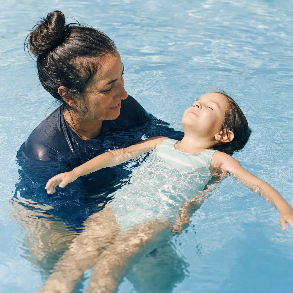 A swim teacher in the water beside a child, close and reassuring, faces lit and warm.